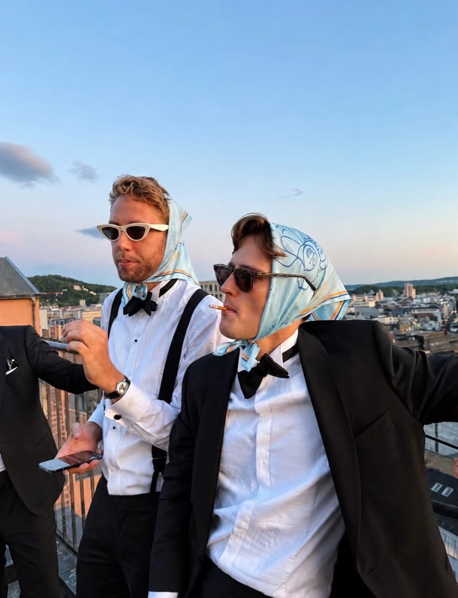 Two men wearing black tuxedo jackets, white shirts, black bow ties, and black suspenders, styled as a costume. One man has a light blue patterned silk scarf wrapped around his head, and the other wears white sunglasses.