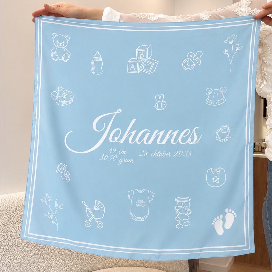 A light blue baby blanket or swaddle held up by a person in a home setting. The blanket features white printed baby-themed icons and the personalized name 'Johannes' in white script.