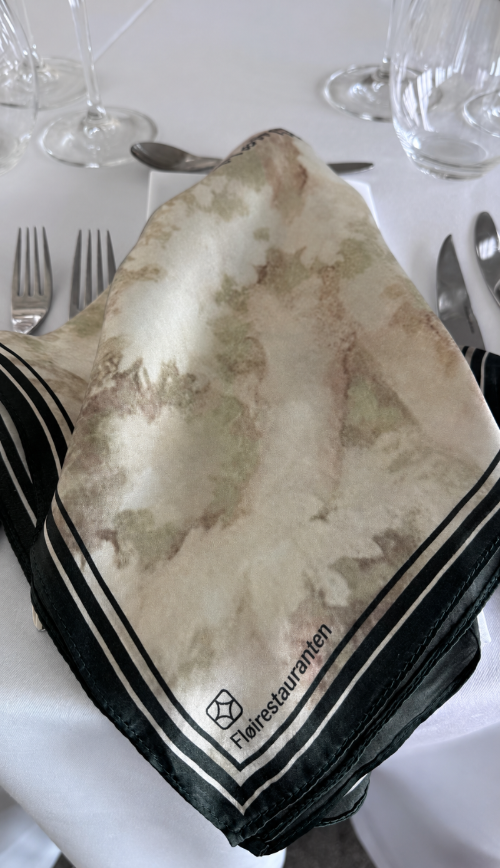 A square scarf with a beige and light green abstract watercolor pattern and black striped borders, displayed on a white tablecloth at a restaurant setting. The brand name 'Fleerrestaurant' is printed on the corner.