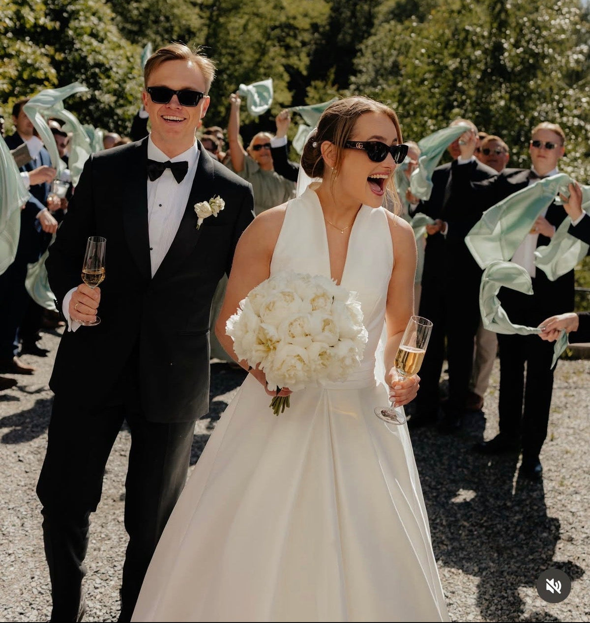 A bride in a white wedding gown and sunglasses walks outdoors with a bouquet, accompanied by a groom in a black tuxedo and sunglasses. Guests wave mint green scarves in the background on a sunny day.
